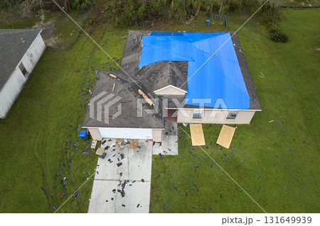 Aerial view of damaged in hurricane Ian house roof covered with blue protective tarp against rain water leaking until replacement of asphalt shingles Aerial view of damaged in hurricane Ian house roof covered with blue protective tarp against rain water leaking until replacement of asphalt shingles 131649939
