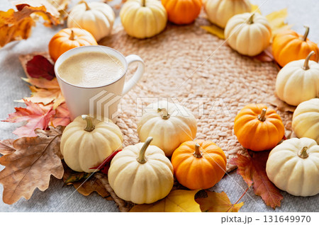 Warm Autumn Morning with Pumpkin Latte. Cup of coffee surrounded by white and orange mini pumpkins with colorful autumn leaves on a woven mat creating a cozy seasonal atmosphere 131649970