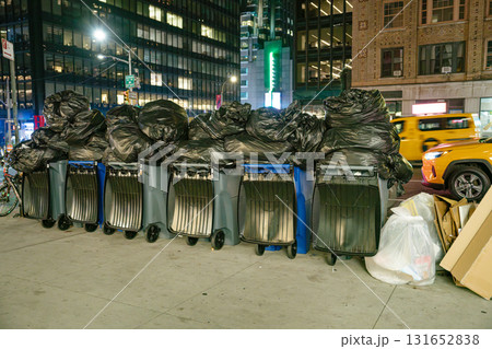 A row of trash bins overloaded with black garbage bags lines the sidewalk in Manhattan. The background reveals busy city streets, lit storefronts, and a yellow taxi passing by at night. 131652838