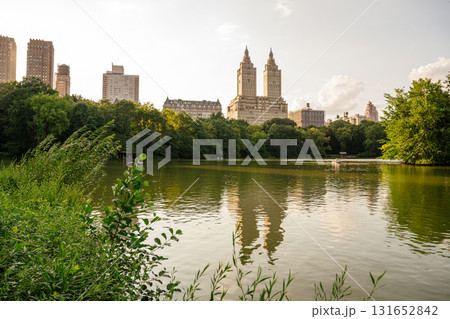 The skyline of Manhattan reflects beautifully in the calm lake of Central Park during golden hour. Lush trees, soft clouds, and peaceful water create a serene urban landscape. The skyline of Manhattan reflects beautifully in the calm lake of Central Park during golden hour. Lush trees, soft clouds, and peaceful water create a serene urban landscape. 131652842