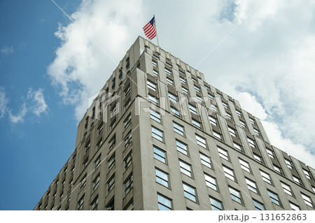 Low-angle view of a beige office building with many square windows and an American flag waving on top. The bright sky with scattered clouds highlights the sharp architectural lines of the structure. 131652863
