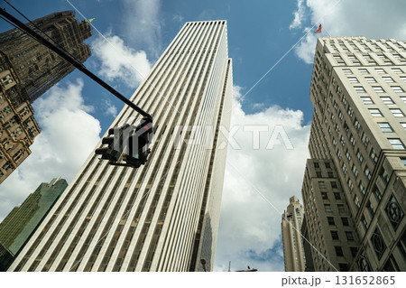 Upward view of Manhattan skyscrapers under a bright blue sky with scattered clouds. A red traffic light is suspended in the foreground, adding contrast to the towering buildings. 131652865