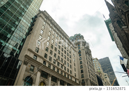 A mix of stone facades and glass buildings rises into a cloudy New York sky. Reflections and textures highlight the contrast between old and new architecture. 131652871