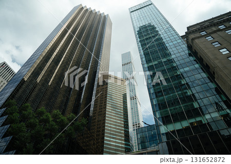 A towering view of glass and steel skyscrapers in Midtown Manhattan, New York City. The architecture reflects light through the cloudy sky, showcasing the vertical power of the urban skyline. 131652872