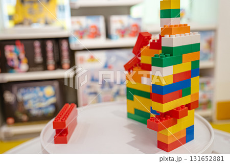 A tall, multicolored constructor tower stands on a round table inside a toy store. The structure is made from red, yellow, blue, green, orange, and white plastic bricks. 131652881