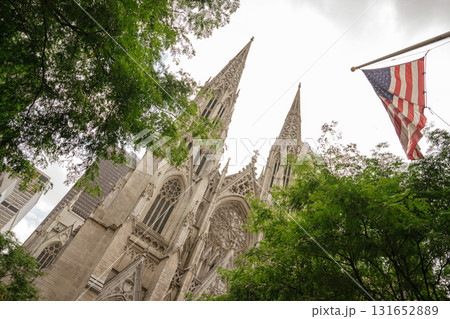 This image shows the iconic twin spires of St. Patrick's Cathedral in New York City, framed by green trees and a waving American flag. The Gothic architecture stands tall among modern skyscrapers. 131652889