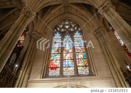 A stunning stained glass window in a gothic-style cathedral, featuring biblical scenes in vivid colors. Tall stone columns and detailed arches frame the sacred artwork, creating a majestic and 131652913