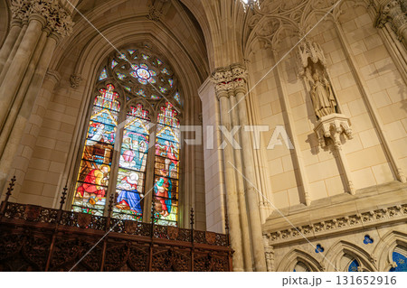 This image showcases a colorful stained-glass window depicting biblical scenes, set within a Gothic arched frame. To the right, a stone statue of a saint is mounted on the cathedral's intricately This image showcases a colorful stained-glass window depicting biblical scenes, set within a Gothic arched frame. To the right, a stone statue of a saint is mounted on the cathedral's intricately 131652916