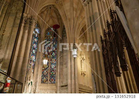 This photo captures a majestic corner of a cathedral with a towering stained-glass window framed by marble columns and vaulted arches. Ornate ironwork, a wall-mounted statue, and soft lighting add to 131652918
