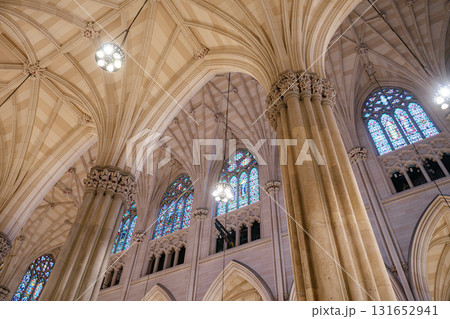 An upward view of a cathedral's vaulted ceiling showcasing intricate Gothic arches and towering columns. Vibrant stained glass windows and elegant chandeliers add beauty and light to the sacred 131652941