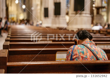 A woman in a colorful shirt is seated alone, leaning forward in silent prayer inside a cathedral. The warm wooden pews and soft lighting create a calm and spiritual atmosphere. A woman in a colorful shirt is seated alone, leaning forward in silent prayer inside a cathedral. The warm wooden pews and soft lighting create a calm and spiritual atmosphere. 131652942