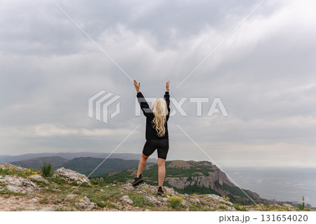 A woman stands on a mountain top, arms raised in the air 131654020