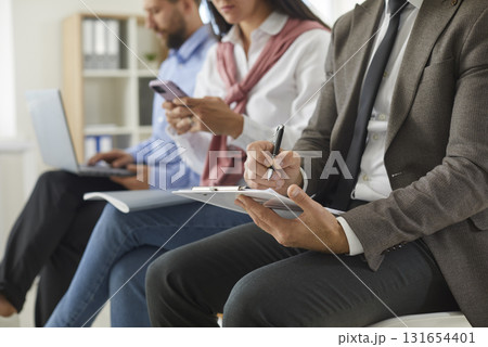 Business man writing on clipboard while others are using laptops and phones in background 131654401