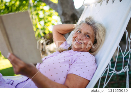 Older woman lying in a hammock and reading book, quiet time in garden. 131655080