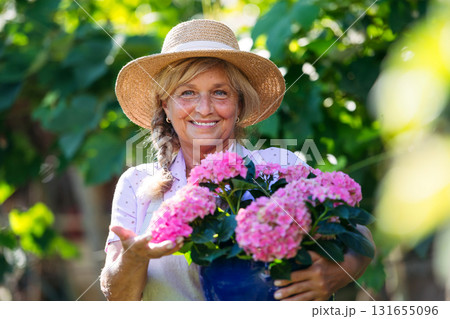 Smiling senior woman holding pot of pink flowers in garden. 131655096