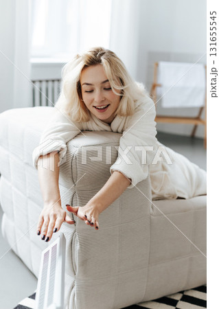 Young woman in a robe warming hands near electric heating radiator indoors 131655545