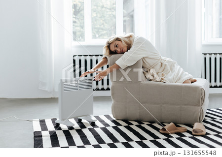 Young woman in a robe warming hands near electric heating radiator indoors 131655548