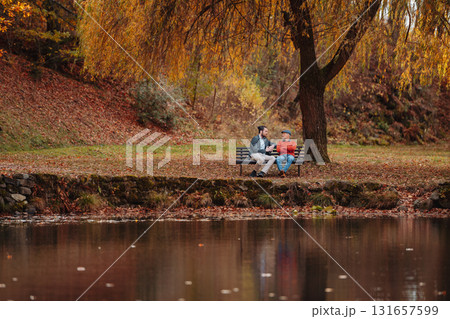 Son and older father sitting on bench by lake and talking. 131657599