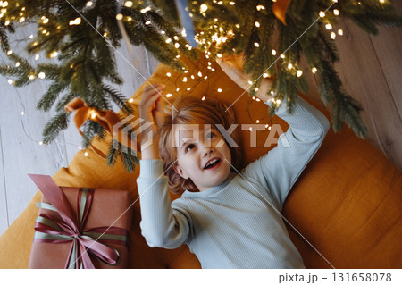 Smiling child top view under Christmas tree with gift box on orange cushion on wooden floor Smiling child top view under Christmas tree with gift box on orange cushion on wooden floor 131658078