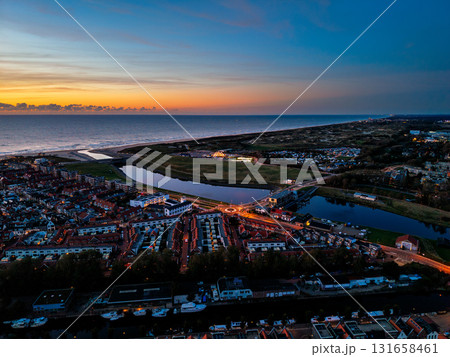 Aerial view of coastal town at sunset dense housing grid, illuminated roundabout, canal flowing to ocean, and vibrant sky over dunes blend urban order with natural serenity. Aerial view of coastal town at sunset dense housing grid, illuminated roundabout, canal flowing to ocean, and vibrant sky over dunes blend urban order with natural serenity. 131658461