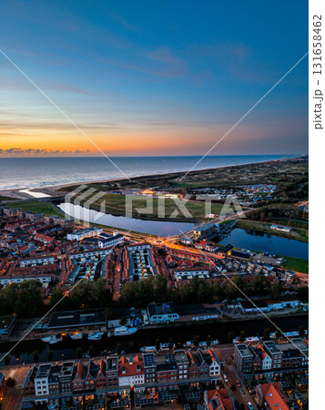 Aerial view of coastal town at sunset dense housing grid, illuminated roundabout, canal flowing to ocean, and vibrant sky over dunes blend urban order with natural serenity. Aerial view of coastal town at sunset dense housing grid, illuminated roundabout, canal flowing to ocean, and vibrant sky over dunes blend urban order with natural serenity. 131658462