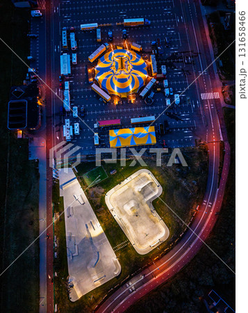 Aerial nighttime view of circus and skatepark blue yellow tent and trailers in parking lot contrast with brightly lit ramps and bowls, blending festive spectacle with modern recreation. 131658466