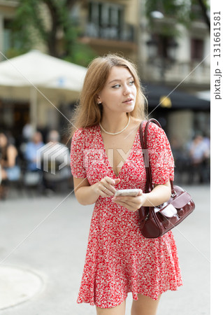 Young woman in summer dress standing on street with phone in hands Young woman in summer dress standing on street with phone in hands 131661581