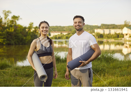 Smiling young man and woman standing on grass, holding yoga mats ready for outdoor workout or yoga. Smiling young man and woman standing on grass, holding yoga mats ready for outdoor workout or yoga. 131661941