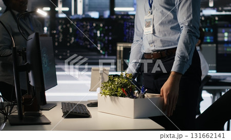 Upset data center worker packing personal items at workstation after being replaced by AI. Programmer removing belongings from server hub workplace desk, fired by artificial intelligence, camera A 131662141
