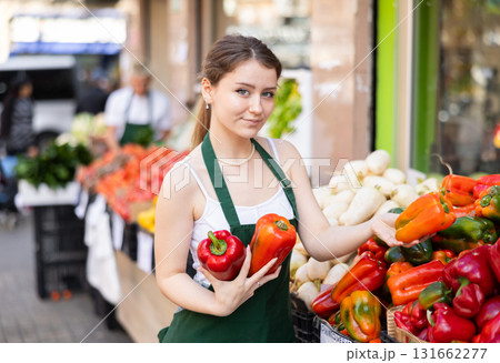 Smiling female saleswoman carefully places ripe bell peppers on a display case at grocery supermarket 131662277