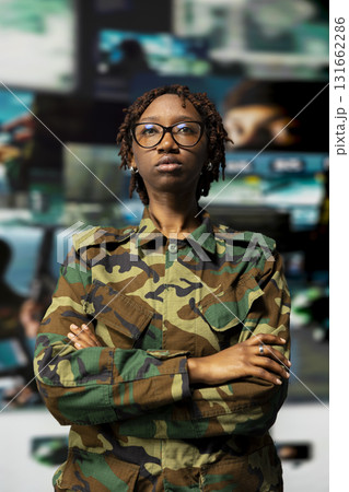 Portrait of officer in camouflage uniform standing with crossed arms against backdrop of digital surveillance screens in military command hub, showing discipline and readiness for defense operations 131662286
