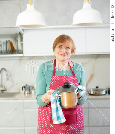Elderly housewife in apron stands in kitchen in front of table and holds stainless pan with lunch 131662412