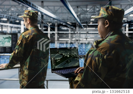 Team of army experts in data center reviewing satellite intelligence feeds on laptop. Military units in server farm command center supporting battlefield strategy with orbital imaging analysis Team of army experts in data center reviewing satellite intelligence feeds on laptop. Military units in server farm command center supporting battlefield strategy with orbital imaging analysis 131662518