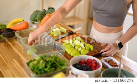 Woman organizing sliced fruits and fresh vegetables into meal containers on a wooden kitchen counter Woman organizing sliced fruits and fresh vegetables into meal containers on a wooden kitchen counter 131666278