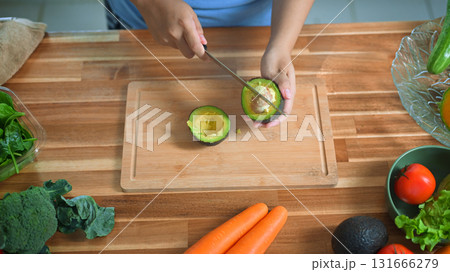 Hands cutting avocado on wooden board surrounded by fresh vegetables, symbolizing healthy eating and clean food 131666279