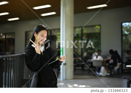 Smiling teenage using a tablet and stylus while leaning on railing in modern school corridor 131666323
