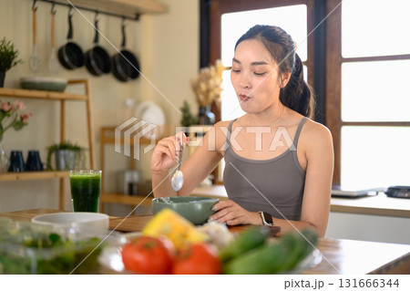 Smiling woman in activewear eating yogurt topped with fresh berries in a bright kitchen 131666344