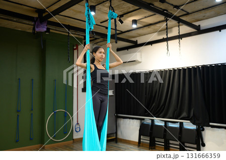 Woman practicing aerial yoga in fitness studio, standing inside blue hammock fabric for balance and strength training Woman practicing aerial yoga in fitness studio, standing inside blue hammock fabric for balance and strength training 131666359