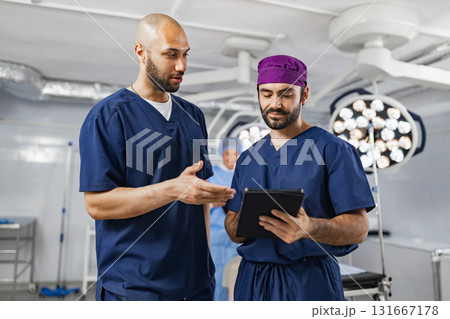Two surgeons in scrubs discuss a patient's medical information in an operating room, using a tablet for reference. Two surgeons in scrubs discuss a patient's medical information in an operating room, using a tablet for reference. 131667178