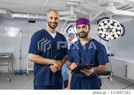 Two surgeons pose in an operating room, with surgical lights and equipment visible in the background, ready for a procedure. Two surgeons pose in an operating room, with surgical lights and equipment visible in the background, ready for a procedure. 131667181