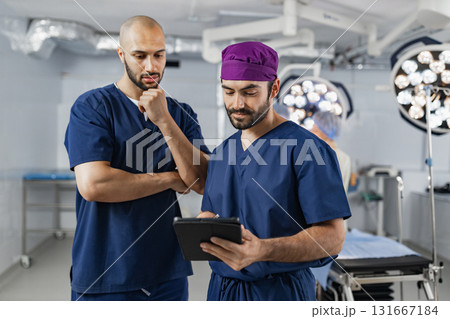 Two surgeons in an operating room review a tablet, likely discussing a patient's medical records before a procedure. 131667184