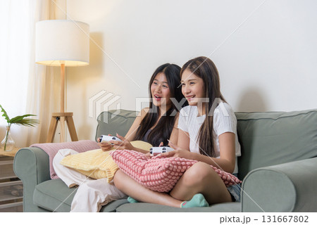 Two young women friends play video game home, teenagers enjoy exciting gameplay, holding controllers, relaxing living room sofa, sharing happy moments. Two young women friends play video game home, teenagers enjoy exciting gameplay, holding controllers, relaxing living room sofa, sharing happy moments. 131667802