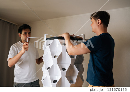 Agricultural technicians assembling indoor vertical hydroponic tower garden, placing top part with grow light onto structure, ready to start growing fresh produce at home. Concept of indoor farming Agricultural technicians assembling indoor vertical hydroponic tower garden, placing top part with grow light onto structure, ready to start growing fresh produce at home. Concept of indoor farming 131670156