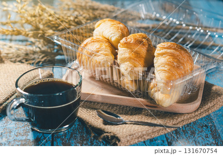 Freshly baked croissants in a plastic container with a hot black coffee cup on a wooden table. Perfect for breakfast or bakery marketing. 131670754