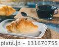 Close-up of a single golden brown croissant on a white plate with a blurry cup of coffee in the background. Shallow depth of field. 131670755