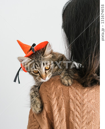 Woman in brown sweater holds tabby kitten wearing small witch's hat on her shoulder. Sweet festive atmosphere of Halloween highlights the bond between humans and animals 131674658