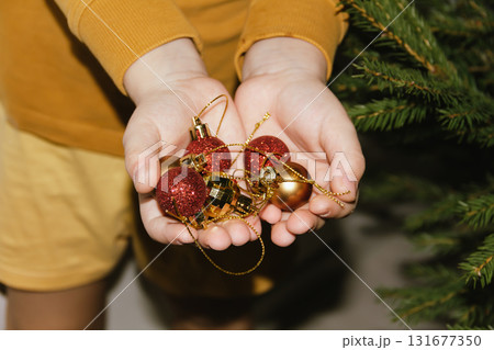 Children's hands hold shiny Christmas tree baubles. The tree is decorated with ornaments, garlands, and lights. Concept for a family Christmas and New Year's celebration. Children's hands hold shiny Christmas tree baubles. The tree is decorated with ornaments, garlands, and lights. Concept for a family Christmas and New Year's celebration. 131677350