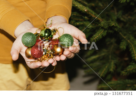 Children's hands hold shiny Christmas tree baubles. The tree is decorated with ornaments, garlands, and lights. Concept for a family Christmas and New Year's celebration. Children's hands hold shiny Christmas tree baubles. The tree is decorated with ornaments, garlands, and lights. Concept for a family Christmas and New Year's celebration. 131677351
