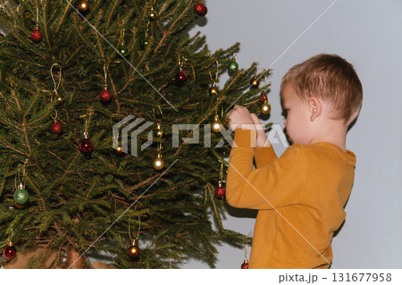 Festive baby boy in yellow sweater getting ready to decorate the Christmas tree, creating a warm holiday atmosphere for celebrating with loving family members tonight. Festive baby boy in yellow sweater getting ready to decorate the Christmas tree, creating a warm holiday atmosphere for celebrating with loving family members tonight. 131677958