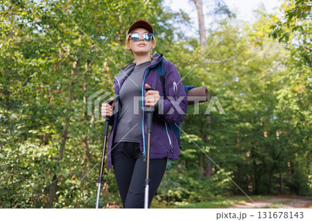 Confident female hiker with trekking poles walking on forest path Confident female hiker with trekking poles walking on forest path 131678143